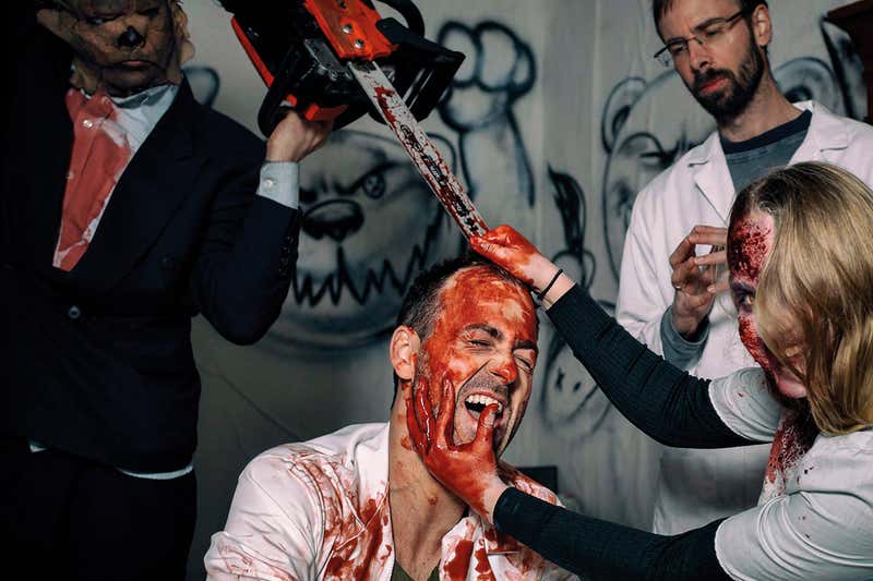 A man covered in fake blood is being scared by another man holding a chainsaw and a woman with a scarred face who's holding him down