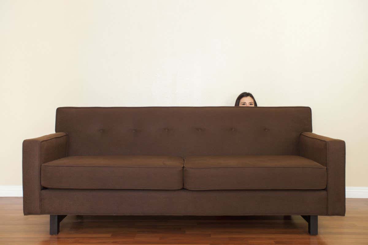 Woman hiding behind sofa against white wall.